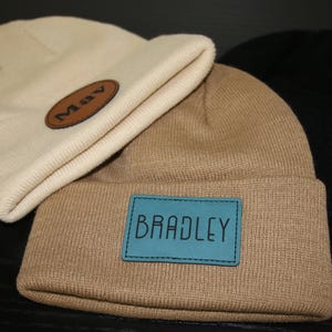 Puede incluir: Dos gorros de punto beige con parches de cuero. Un parche dice "MAY" y el otro dice "BRADLEY".