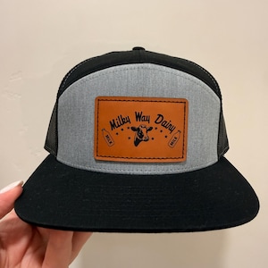 Op de afbeelding: Een zwarte en grijze truckerpet met een zwarte klep en mesh achterkant. De voorkant heeft een grijs paneel met een bruine leren patch. De patch leest "Milky Way Dairy" met een koeienafbeelding en melkfles-iconen.
