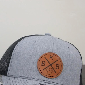 May include: Gray and black trucker hat with a leather patch. The patch features the letters "BKB" in a cross design, with a Mickey Mouse head silhouette. The hat has a Richardson 112 tag.