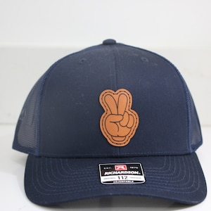 May include: A navy blue trucker hat with a brown leather peace sign patch. The hat is adjustable and has a mesh back.