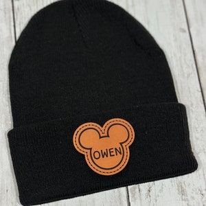 Puede incluir: Gorro de punto negro con un parche de cuero marrón en forma de oreja de Mickey Mouse que dice "OWEN".