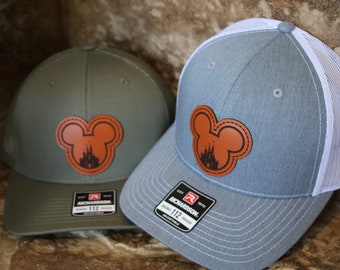 May include: Two baseball caps with a brown leather Mickey Mouse patch featuring the silhouette of Cinderella's castle. The cap on the left is olive green with a black mesh back. The cap on the right is light blue with a white mesh back.