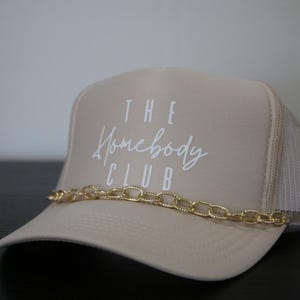 Może przedstawiać: Beżowa czapka trucker z haftowanym białym logo z napisem "THE Homebody CLUB". Złoty łańcuch owinięty wok&oacute;ł czapki.