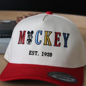 May include: White baseball cap with a red brim. The front of the cap has colourful embroidered text that says "MICKEY EST. 1928".