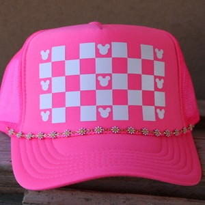 Puede incluir: Una gorra de camionero rosa ne&oacute;n con un patr&oacute;n de cuadros blancos en el panel frontal. El patr&oacute;n incluye peque&ntilde;as siluetas de cabezas de rat&oacute;n blancas integradas en la cuadr&iacute;cula. Una fina cadena de peque&ntilde;as margaritas blancas y amarillas adorna la visera.