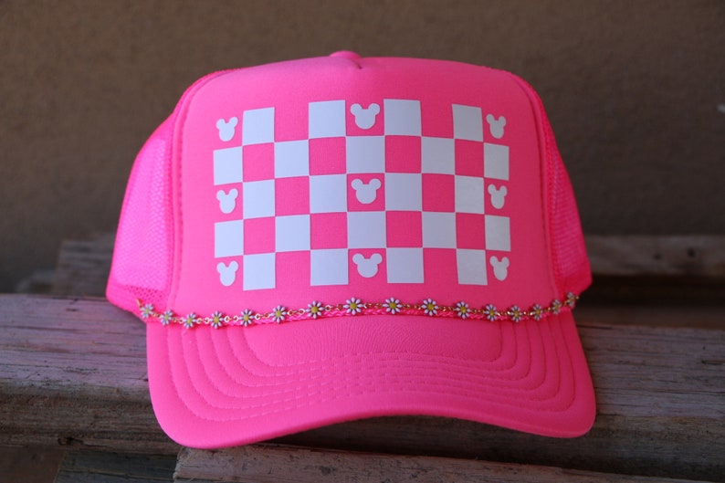 Puede incluir: Una gorra de camionero rosa ne&oacute;n con un patr&oacute;n de cuadros blancos en el panel frontal. El patr&oacute;n incluye peque&ntilde;as siluetas de cabezas de rat&oacute;n blancas integradas en la cuadr&iacute;cula. Una fina cadena de peque&ntilde;as margaritas blancas y amarillas adorna la visera.