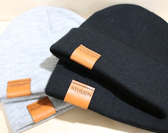 May include: Two knit beanies with leather patches. One beanie is gray and the other is black. Both beanies have a brown leather patch with the word "ATHLON" printed on them.
