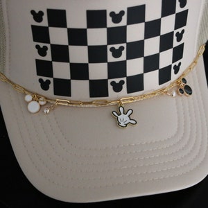 Puede incluir: Una gorra tipo trucker beige con un patr&oacute;n de cuadros en blanco y negro y peque&ntilde;as siluetas de cabeza de rat&oacute;n. Una cadena dorada est&aacute; sujeta al frente, adornada con dijes de esmalte que incluyen una cabeza de rat&oacute;n blanca, un guante y c&iacute;rculos.