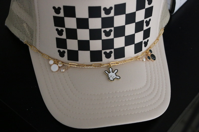 Puede incluir: Una gorra tipo trucker beige con un patr&oacute;n de cuadros en blanco y negro y peque&ntilde;as siluetas de cabeza de rat&oacute;n. Una cadena dorada est&aacute; sujeta al frente, adornada con dijes de esmalte que incluyen una cabeza de rat&oacute;n blanca, un guante y c&iacute;rculos.