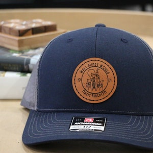 Peut inclure: Casquette de camionneur bleu marine avec un dos en filet gris et une visière incurvée. Un écusson rond en cuir affiche "Walt Disney World Magic Kingdom" et un dessin de château. La casquette est réglable.