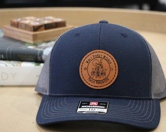 May include: Navy blue trucker hat with a gray mesh back and a curved brim. A circular leather patch features the words "Walt Disney World Magic Kingdom" and a castle graphic. The hat is adjustable.