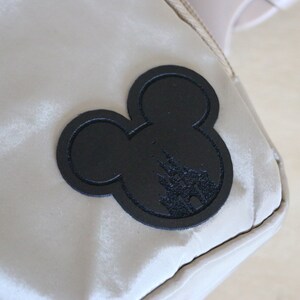 K&ouml;nnte beinhalten: Eine beige Tasche mit einem schwarzen Mickey-Mouse-Silhouetten-Patch, der die Silhouette von Schloss Aschenputtel zeigt.