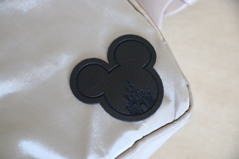 K&ouml;nnte beinhalten: Eine beige Tasche mit einem schwarzen Mickey-Mouse-Silhouetten-Patch, der die Silhouette von Schloss Aschenputtel zeigt.