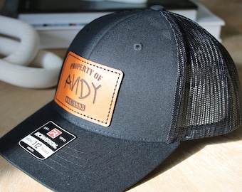 May include: A black trucker hat featuring a tan rectangular leather patch on the front. The patch is engraved with the text PROPERTY OF ANDY EST. 1995. The hat has a black mesh back and a black brim with a Richardson 112 Youth sticker attached.