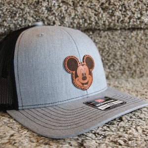 Op de afbeelding: Een grijze en zwarte truckercap met een bruine leren Mickey Mouse-patch op de voorkant. De pet heeft een zwart mesh-achterkant en een grijze klep.