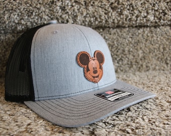 May include: A gray and black trucker hat with a brown leather Mickey Mouse patch on the front. The hat has a black mesh back and a gray brim.