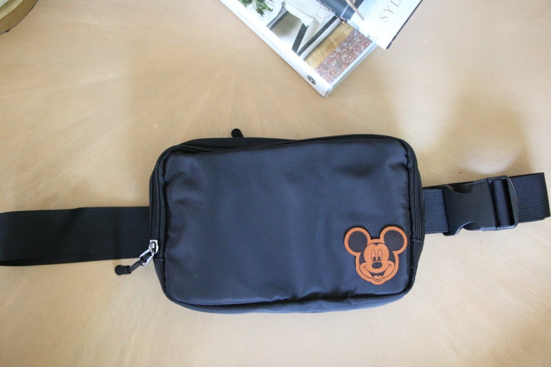 K&ouml;nnte beinhalten: Eine schwarze Bauchtasche mit einem braunen Mickey Mouse-Gesichts-Patch. Die Tasche hat einen schwarzen verstellbaren Gurt mit einer Schnalle.