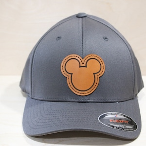 May include: A gray baseball cap with a brown leather Mickey Mouse head patch. The cap has a black Flexfit label on the back.