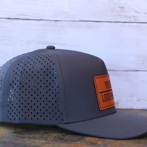 May include: A dark gray baseball cap featuring a perforated back panel for ventilation and a solid front panel. A rectangular tan leather-like patch is stitched onto the front, displaying the black text "YOUR LOGO HERE" with a horizontal line.