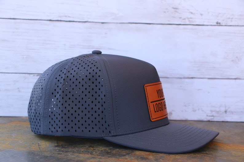 May include: A dark gray baseball cap featuring a perforated back panel for ventilation and a solid front panel. A rectangular tan leather-like patch is stitched onto the front, displaying the black text "YOUR LOGO HERE" with a horizontal line.