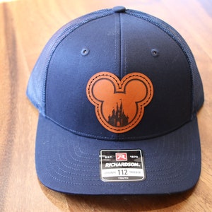 Peut inclure: Une casquette de baseball bleu marine pour enfant avec un patch en cuir marron représentant une silhouette de Mickey Mouse avec le château de Cendrillon à l'intérieur. Le patch est cousu sur les bords. La casquette a un dos en mesh et une étiquette noire avec le texte "EST. 1970 RICHARDSON. ORIGINAL 112 TRUCKER YOUTH".