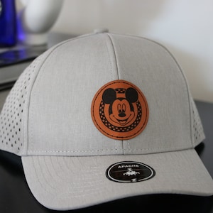 May include: A gray baseball cap with a brown leather patch featuring Mickey Mouse. The patch has a black outline and white polka dots. The cap has a mesh back and the word "APACHE" is printed on the back.