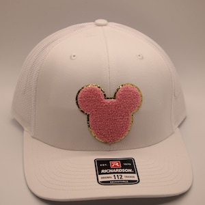 Może przedstawiać: Biała czapka trucker z nadrukiem Myszki Miki w kolorze różowym i złotym. Czapka ma siateczkowe plecy i regulowany pasek z logo Richardson 112.