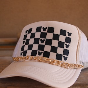 Puede incluir: Una gorra tipo trucker de color crema con malla blanca en la parte posterior y un panel frontal con un patr&oacute;n de tablero de ajedrez negro y crema. Peque&ntilde;as siluetas de orejas de rat&oacute;n negras est&aacute;n integradas en la cuadr&iacute;cula. Una cadena dorada cruza la visera.