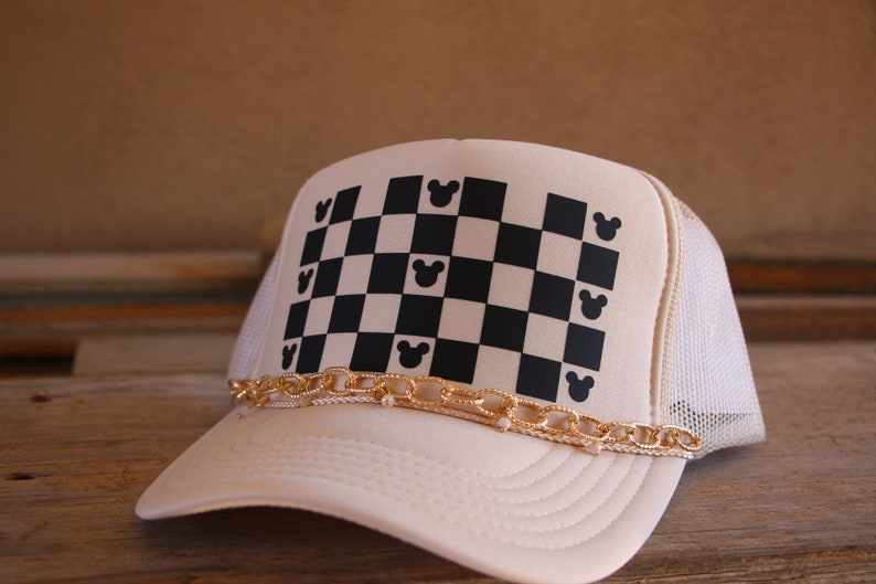 Puede incluir: Una gorra tipo trucker de color crema con malla blanca en la parte posterior y un panel frontal con un patr&oacute;n de tablero de ajedrez negro y crema. Peque&ntilde;as siluetas de orejas de rat&oacute;n negras est&aacute;n integradas en la cuadr&iacute;cula. Una cadena dorada cruza la visera.