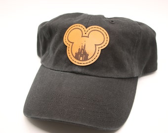 May include: A black baseball cap with a brown leather Mickey Mouse ear patch featuring a silhouette of Cinderella's Castle.