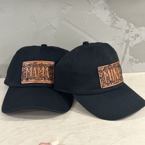 Op de afbeelding: Twee zwarte baseballpetten met bruine leren patches. De patch op de linker pet zegt "MAMA" en de patch op de rechter pet zegt "MINE".