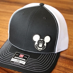Könnte beinhalten: Schwarz-weiße Trucker-Kappe mit weißem Netzrücken. Die Kappe hat einen schwarz-weißen Mickey-Mouse-Aufnäher auf der Vorderseite. Die Kappe ist verstellbar und hat ein weißes Nähdetail am Rand.
