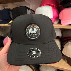 May include: A black baseball cap featuring a circular patch on the front with the text Quality Ornamental Iron Est 1985 and an illustration of a welder. A round sticker on the brim displays a helicopter graphic and the word Blackhawk.