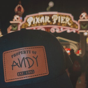 May include: A black and brown baseball cap with a brown leather patch that says "Property of Andy Est. 1995". The cap is in front of a brightly lit sign that says "Pixar Pier".