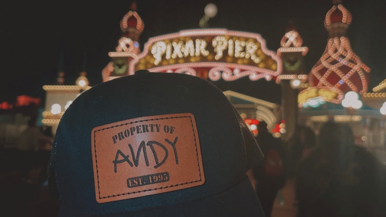 May include: A black and brown baseball cap with a brown leather patch that says "Property of Andy Est. 1995". The cap is in front of a brightly lit sign that says "Pixar Pier".