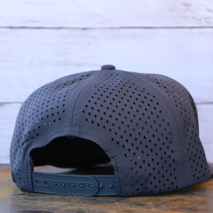 May include: A dark gray baseball cap featuring a perforated crown for ventilation. The back of the cap includes an adjustable plastic snapback closure with a matching dark gray strap and button details, showcasing a lightweight, breathable design.
