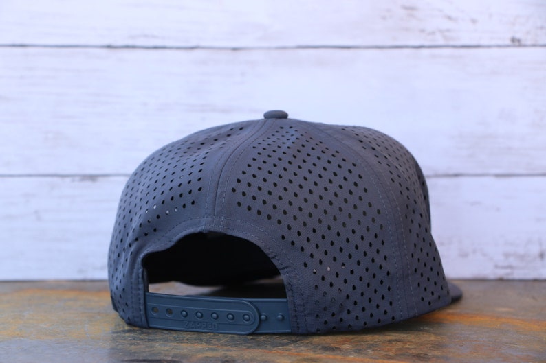 May include: A dark gray baseball cap featuring a perforated crown for ventilation. The back of the cap includes an adjustable plastic snapback closure with a matching dark gray strap and button details, showcasing a lightweight, breathable design.