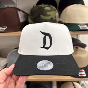 Pode incluir: Boné de beisebol branco e preto com uma letra "D" preta bordada na frente. O boné tem uma aba plana e um adesivo "Cal Hat". Outros bonés são visíveis nas prateleiras atrás do boné principal.