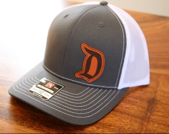 May include: A gray and white trucker hat with a brown leather 'D' embroidered on the front. The hat has a mesh back and a curved brim.