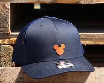 May include: Navy blue trucker hat with a mesh back and a curved brim. The hat features a brown Mickey Mouse head emblem on the front. The hat is adjustable and has a Richardson 112 tag.