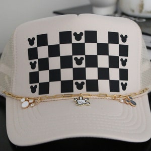 Peut inclure: Casquette de camionneur beige avec un motif à carreaux noir et blanc sur le devant, orné de silhouettes de Mickey Mouse. Une chaîne dorée avec des breloques orne la visière. Le dos est en mesh.