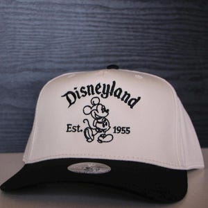 May include: White and black baseball cap with a black brim. The front of the cap features a black embroidered logo that says "Disneyland Est. 1955" with a cartoon image of Mickey Mouse.