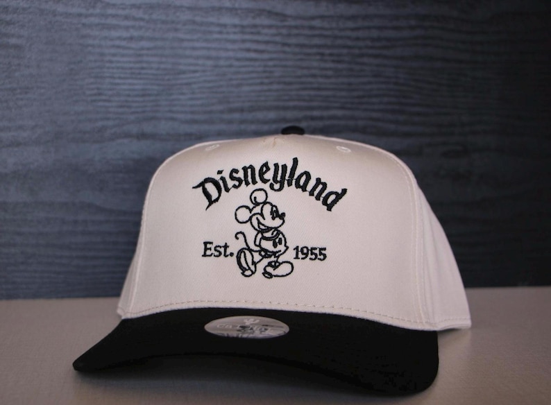 K&ouml;nnte beinhalten: Wei&szlig;-schwarze Baseballkappe mit schwarzem Schirm. Die Vorderseite der Kappe zeigt ein schwarzes gesticktes Logo mit der Aufschrift "Disneyland Est. 1955" mit einem Cartoon-Bild von Mickey Mouse.