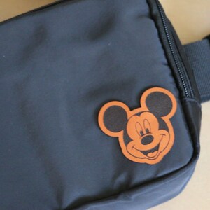 K&ouml;nnte beinhalten: Eine schwarze Bauchtasche mit einem braunen Leder-Mickey-Mouse-Aufn&auml;her auf der Vorderseite. Die Bauchtasche hat einen schwarzen verstellbaren Gurt.