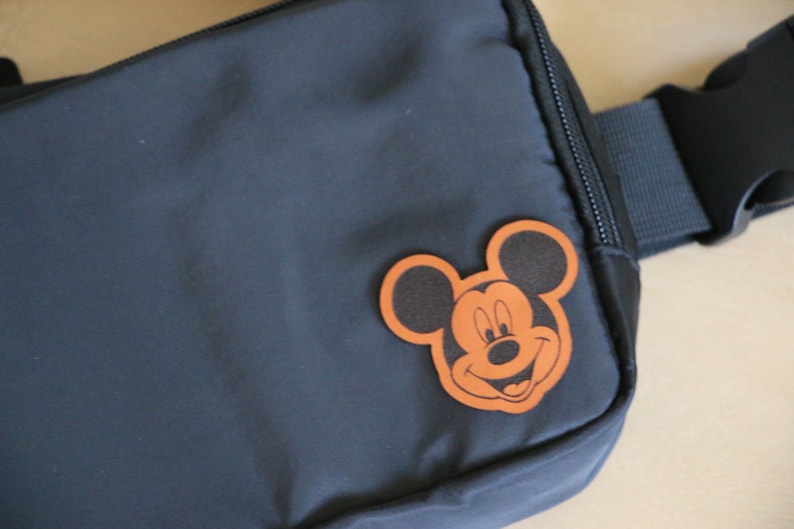 K&ouml;nnte beinhalten: Eine schwarze Bauchtasche mit einem braunen Leder-Mickey-Mouse-Aufn&auml;her auf der Vorderseite. Die Bauchtasche hat einen schwarzen verstellbaren Gurt.