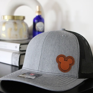 May include: A gray and black trucker hat with a brown leather Mickey Mouse head patch.