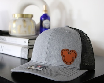 May include: A gray and black trucker hat with a brown leather Mickey Mouse head patch.