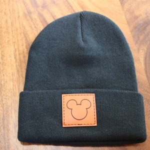 Peut inclure: Un bonnet noir avec un patch en cuir marron représentant une silhouette de Mickey Mouse.