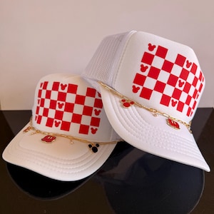May include: Two white trucker hats with red and white checkered patterns and Mickey Mouse silhouettes. Each hat features a gold chain with Mickey Mouse charms. One hat has a black Mickey Mouse charm, and the other has a red Mickey Mouse charm.