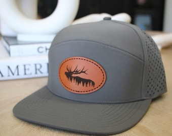 Puede incluir: Una gorra de béisbol gris y marrón con un parche de cuero que presenta una silueta de un alce. La gorra tiene un panel trasero de malla.